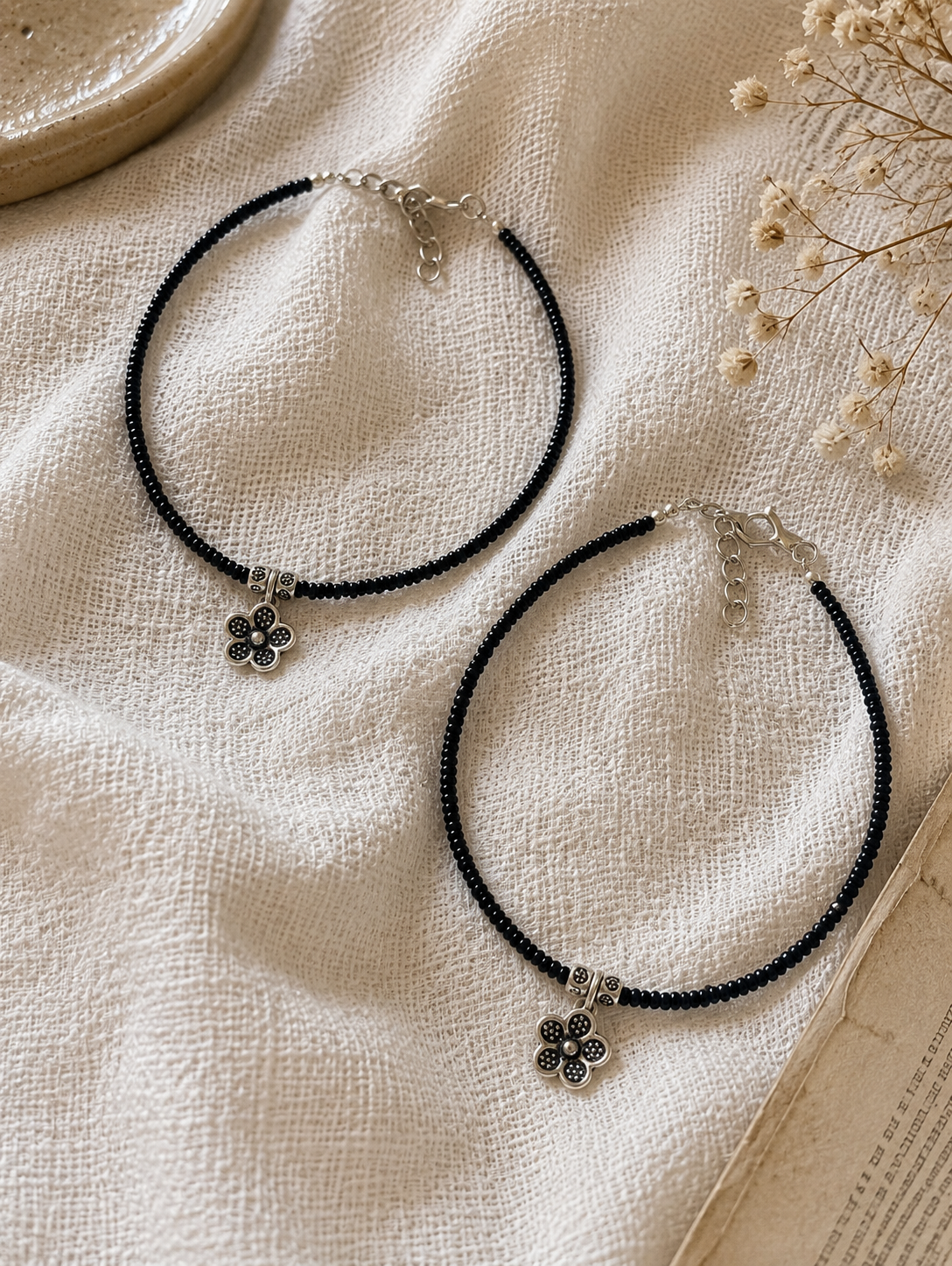 Two black beaded anklets with silver flower pendants on a textured beige surface.
