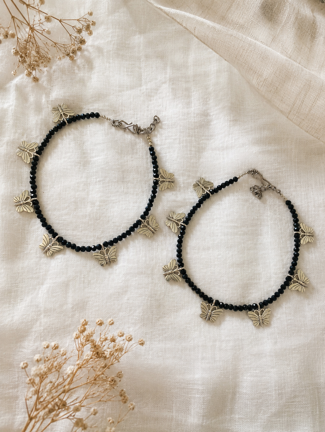 Black beaded Anklets with butterfly charms on a light fabric background