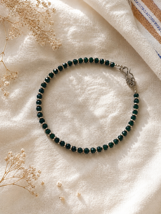 Green beaded anklet on a soft white fabric background with dried flowers.