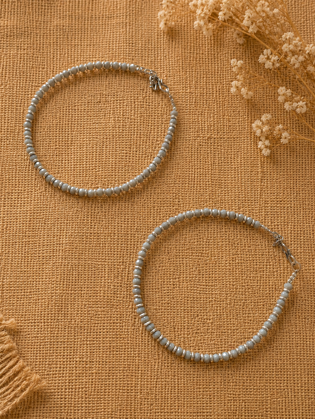 Two beaded anklets on a textured brown surface with dried flowers.