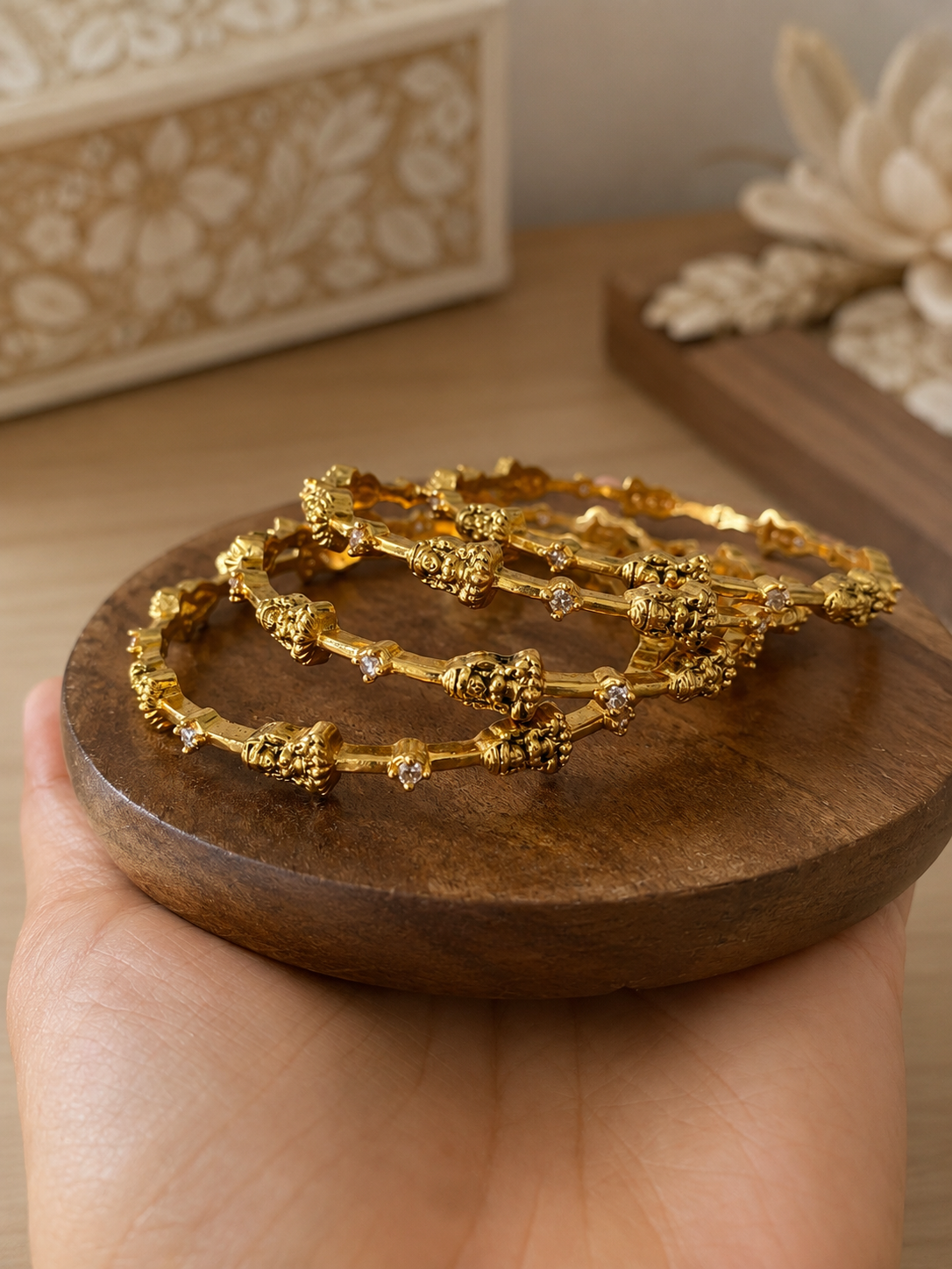 Gold bangles on a wooden tray held by a hand with a decorative background