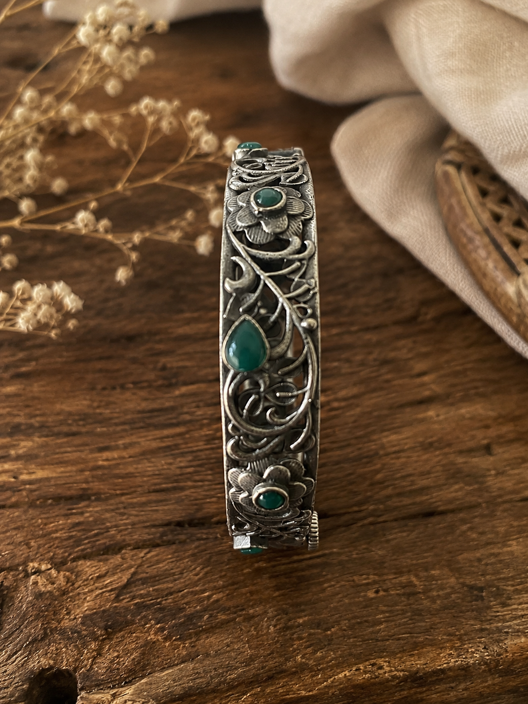 Silver bracelet with green stones on a wooden surface with dried flowers and fabric.