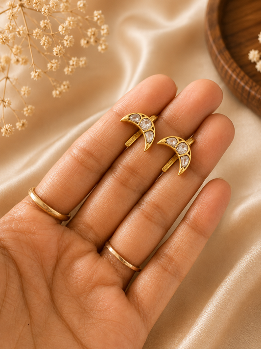 Close-up of a hand presenting a pair of gold-toned crescent moon ear cuffs embedded with clear, traditional white Jadau Kundan stones against a neutral satin backdrop.