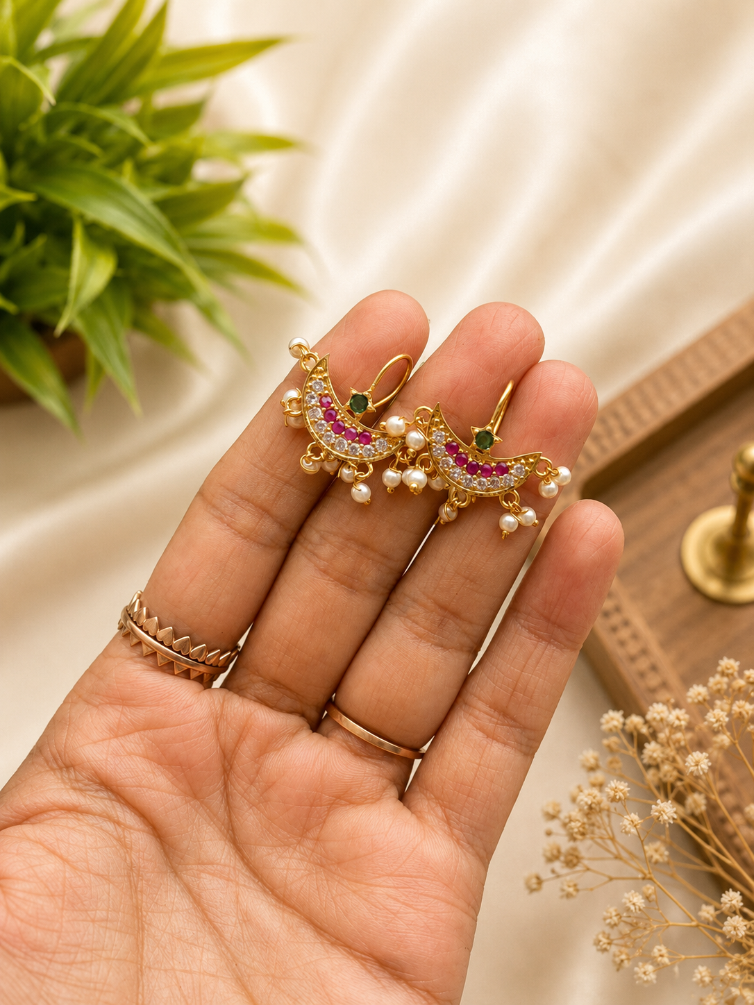 Traditional gold-plated moon-shaped Bugudi earrings featuring ruby and emerald stones, white cz accents, and hanging seed pearls, held in a person's hand against a soft silk background.