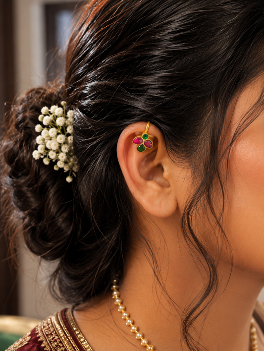 Close-up of a bride with an elegant bun hairstyle decorated with white baby's breath flowers, wearing a traditional gold-plated Leaf Style Jadau Bugudi ear cuff featuring ruby pink and green emerald stones on the upper cartilage.