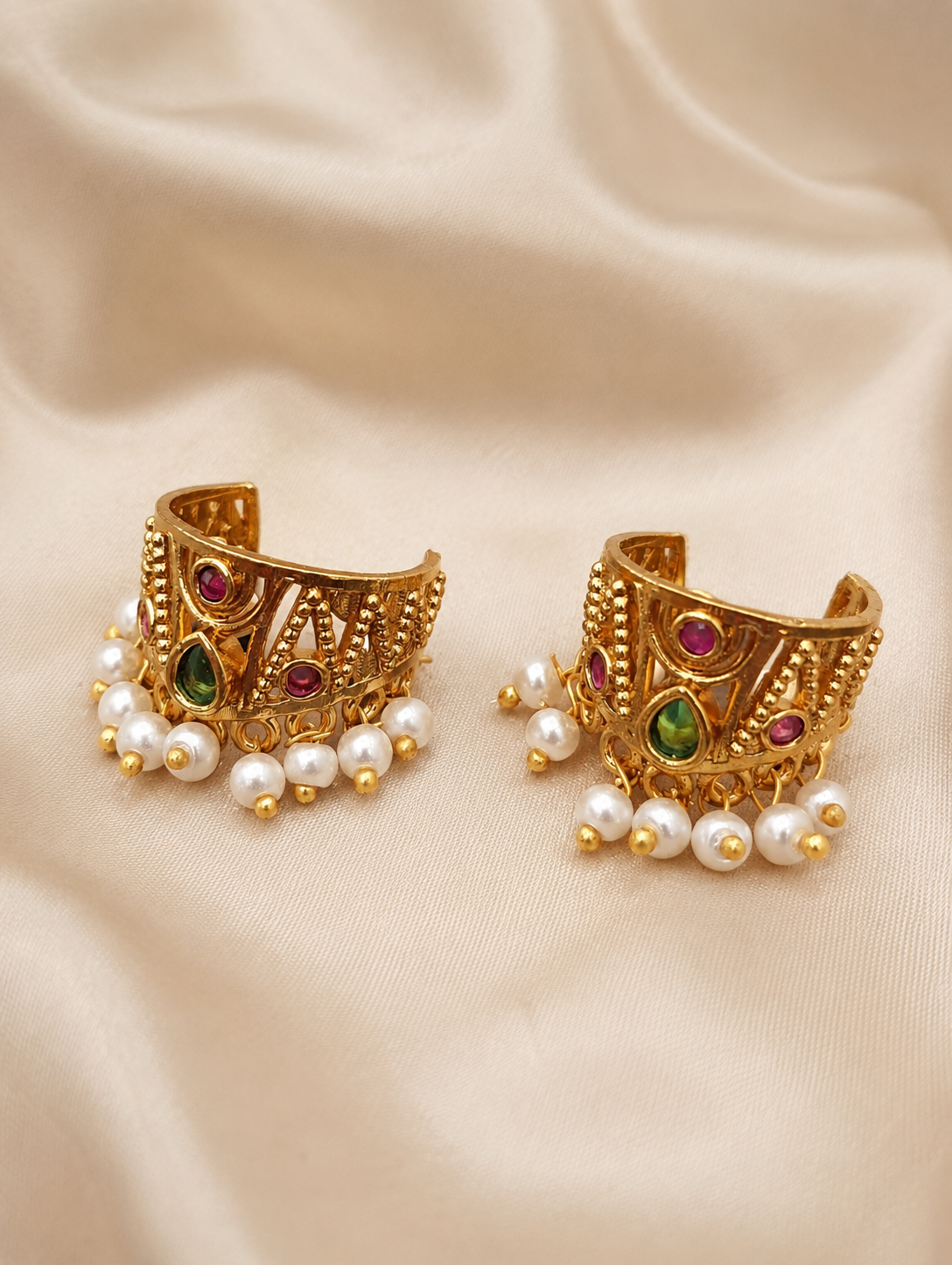 A pair of traditional Indian gold-plated Bugudi earrings for the upper ear cartilage, featuring intricate gold filigree, embedded ruby-red and teardrop green kemp stones, and a fringe of delicate white pearls at the bottom, displayed on a soft, cream-colored satin fabric.