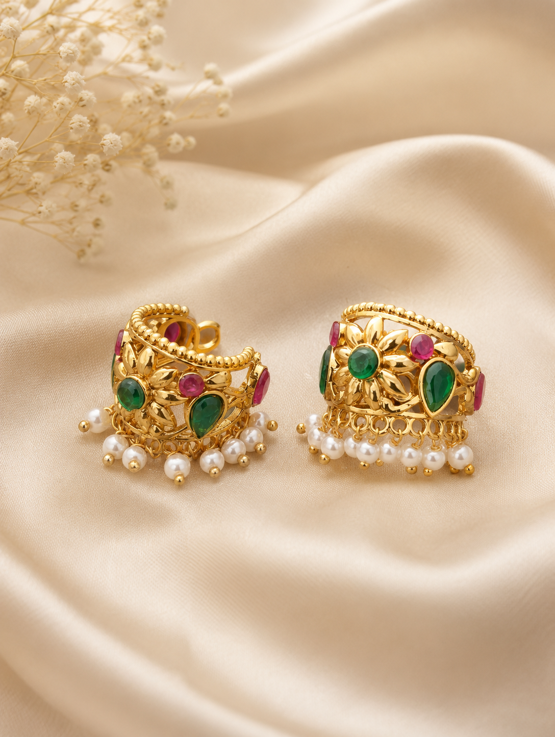A pair of traditional gold-plated Bugudi cartilage earrings featuring a sculpted gold floral motif with a center emerald-green stone, accent ruby-red and green gems, and a fringe of pearl drops against champagne-colored silk fabric.