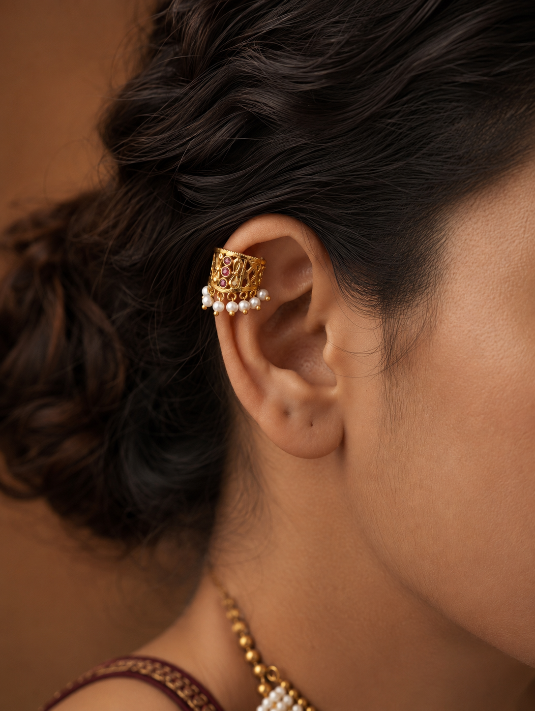 A pair of traditional gold-plated Indian Bugudi cartilage earrings featuring a geometric gold cutwork band adorned with a vertical row of three round ruby-red kemp stones and a bottom trim of white pearl drops, set against a draped off-white linen background.