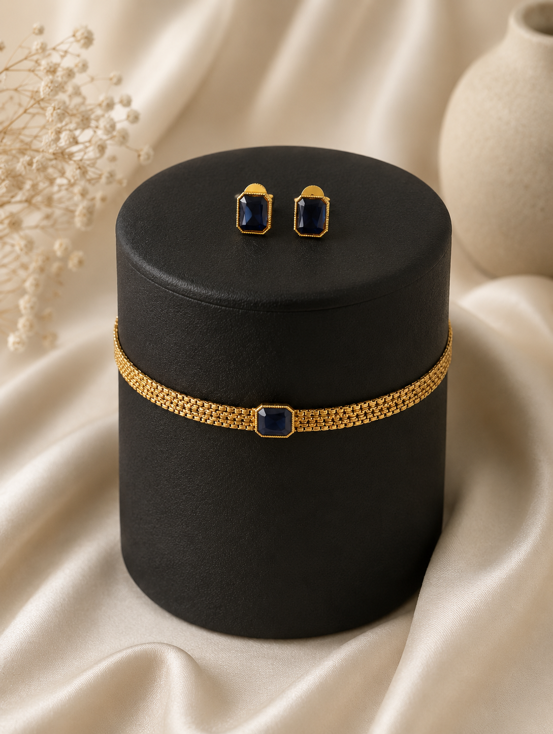 Neelam minimalist jewelry set showcasing a gold-toned mesh choker necklace with a central emerald-cut blue sapphire stone, wrapped around a black cylindrical display stand with matching sapphire stud earrings.