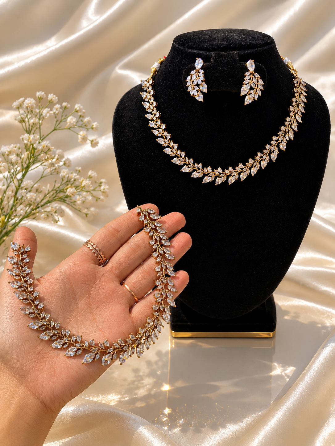 A beautiful gold-plated jewelry set shown draped on a hand and displayed on a black jewelry bust, featuring a leafy vine collar necklace and matching drop earrings entirely embedded with clear marquise-cut CZ stones against a shiny cream satin background.