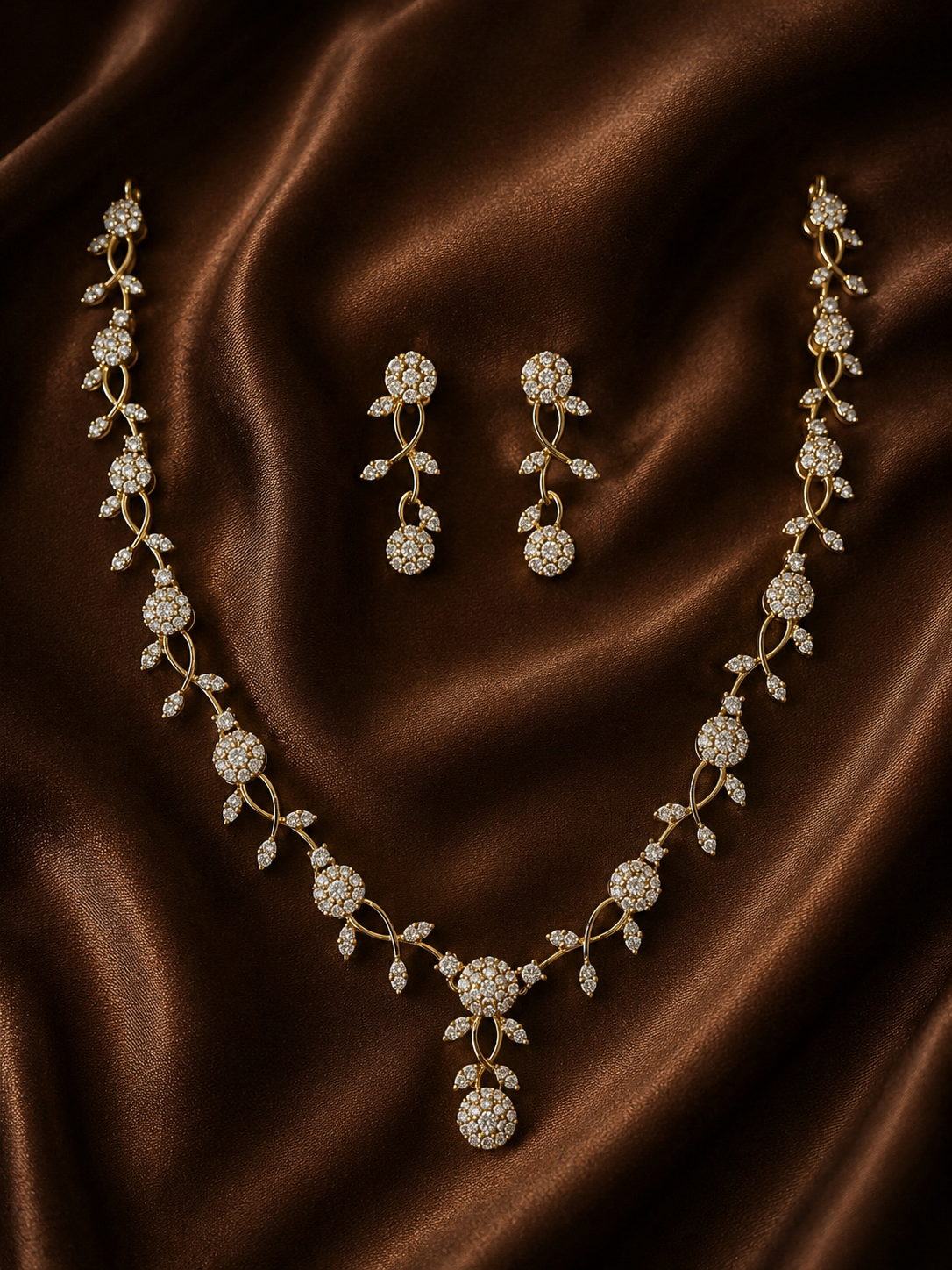 A gold-plated necklace and matching drop earrings set with a delicate interwoven vine design and round, crystal-studded floral motifs, resting on a draped brown satin fabric.