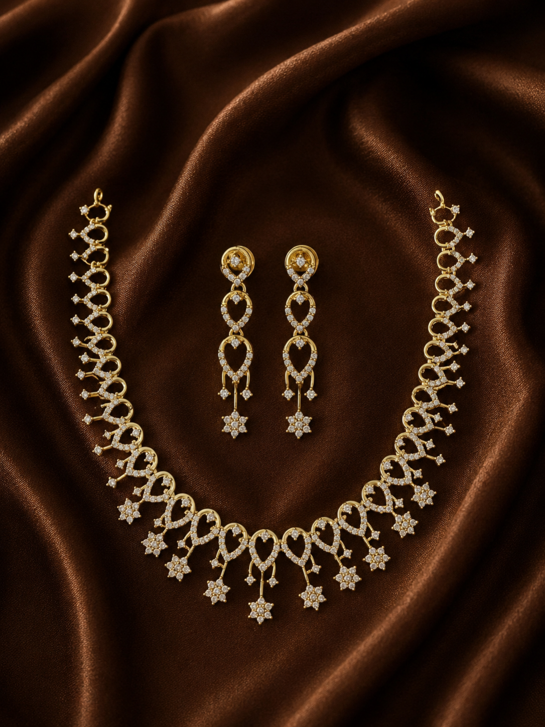 A luxury gold-plated necklace and matching long drop earrings set, detailed with crystal-studded loops and star-shaped dangles, displayed on a rich, draped brown satin background.