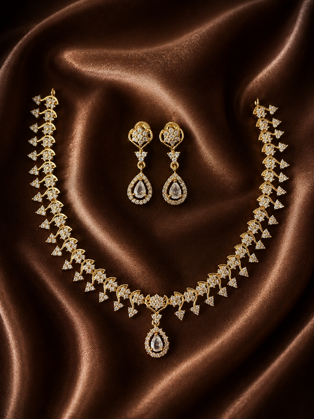 A gold-plated bridal necklace and matching long drop earrings set, detailed with small crystal accents and prominent crystal teardrop charms, laid out on a shimmering brown satin background.