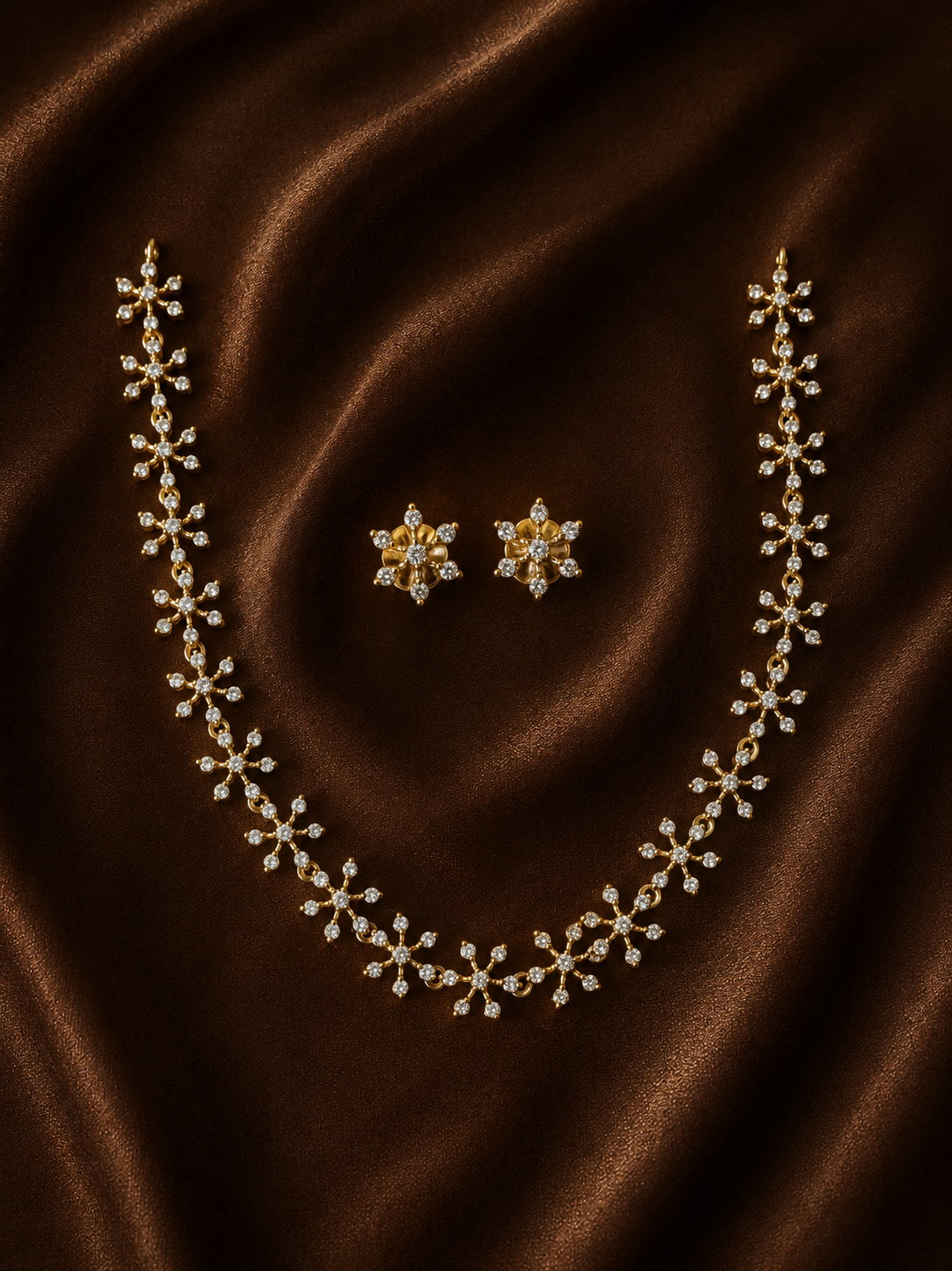 A gold-plated necklace chain and matching stud earrings set, featuring brilliant crystal-studded starburst and flower designs, arranged on a rich, draped brown satin fabric background.