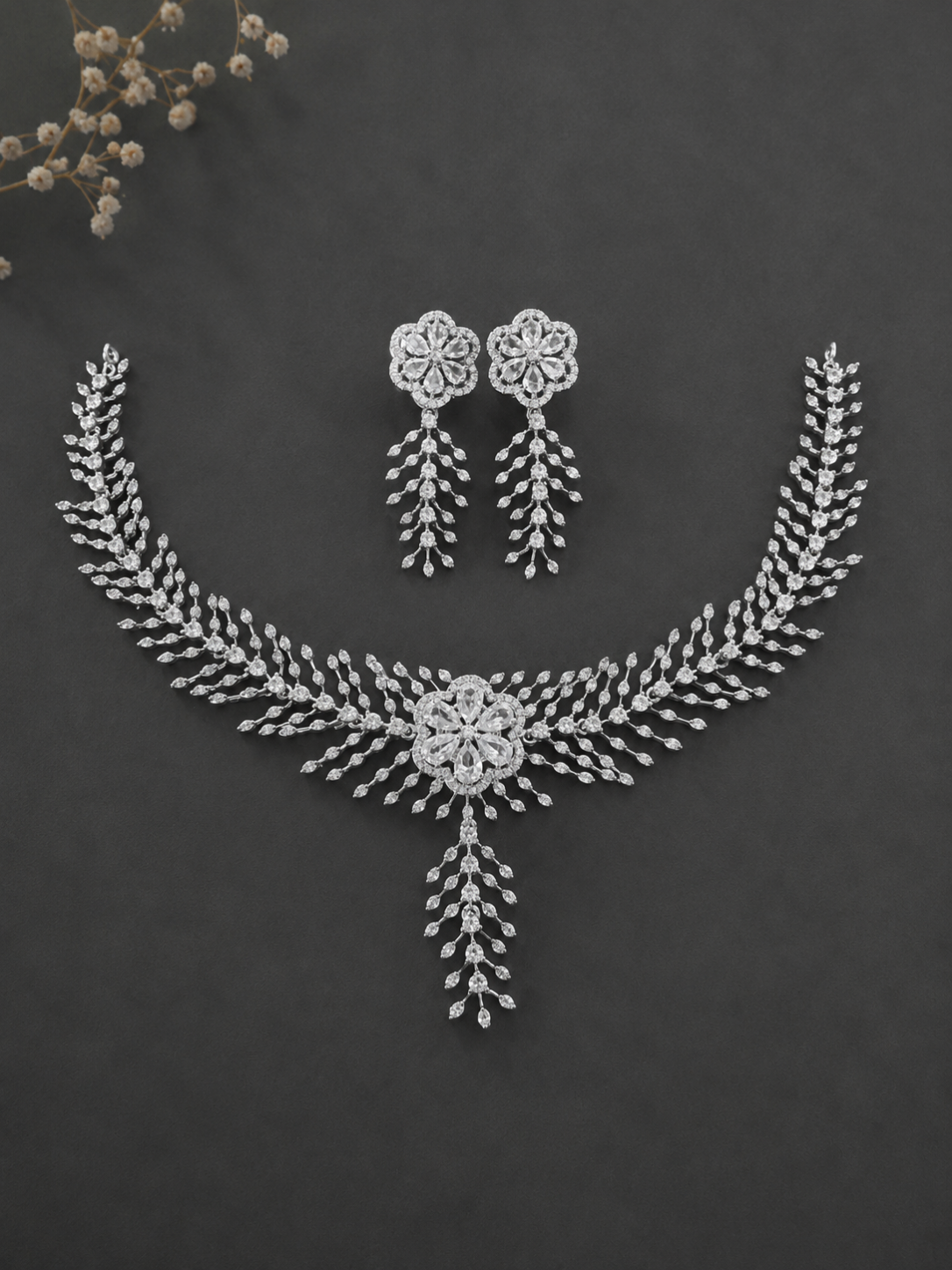 A premium silver-toned cubic zirconia jewelry set featuring a detailed floral centerpiece necklace with a hanging leaf tendril and matching dangle earrings on a dark grey background.