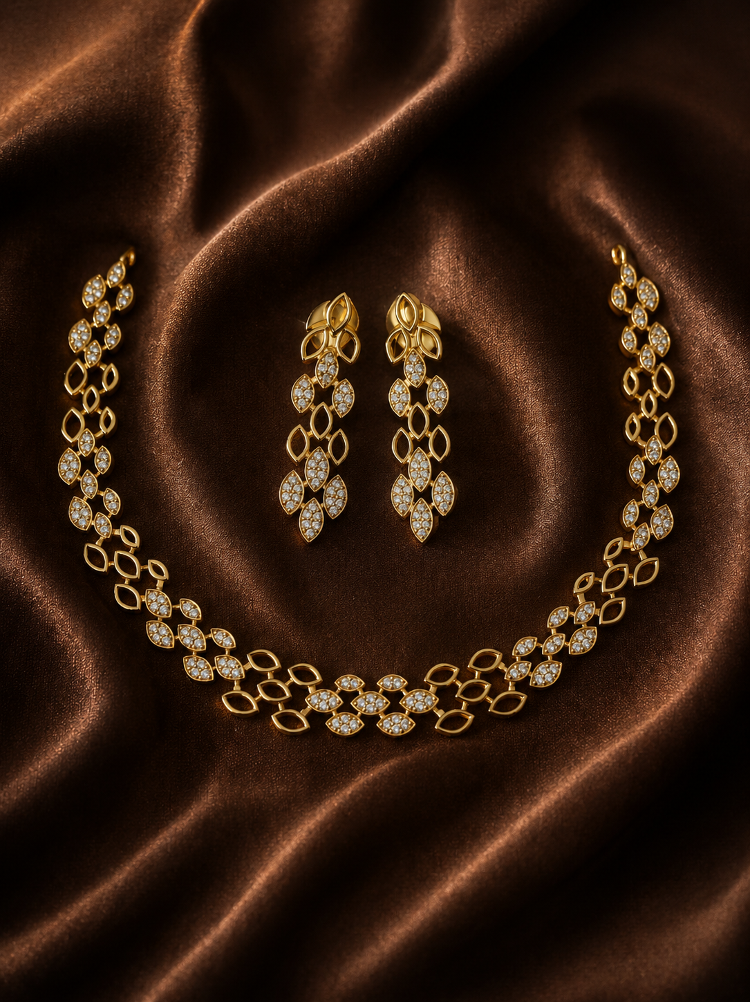 A luxury gold jewelry set including a collar necklace and matching dangle earrings with diamond-encrusted leaf patterns, displayed on a brown satin fabric background.