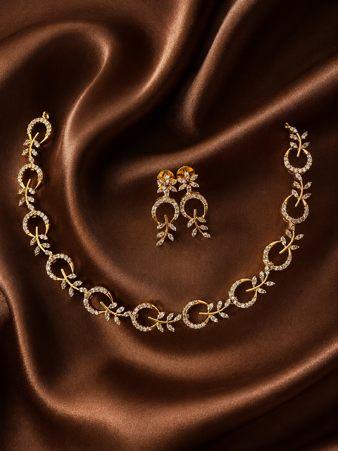 A gold-plated choker necklace and matching short drop earrings featuring an intertwined circle and crystal leaf sprig design, laid flat on glossy brown satin.