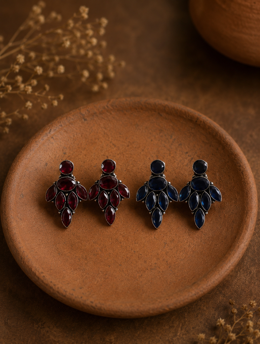 Two pairs of Uma oxidized silver cluster earrings, one set featuring ruby red navette gemstones and the other featuring deep sapphire blue gemstones, displayed on a small clay dish.