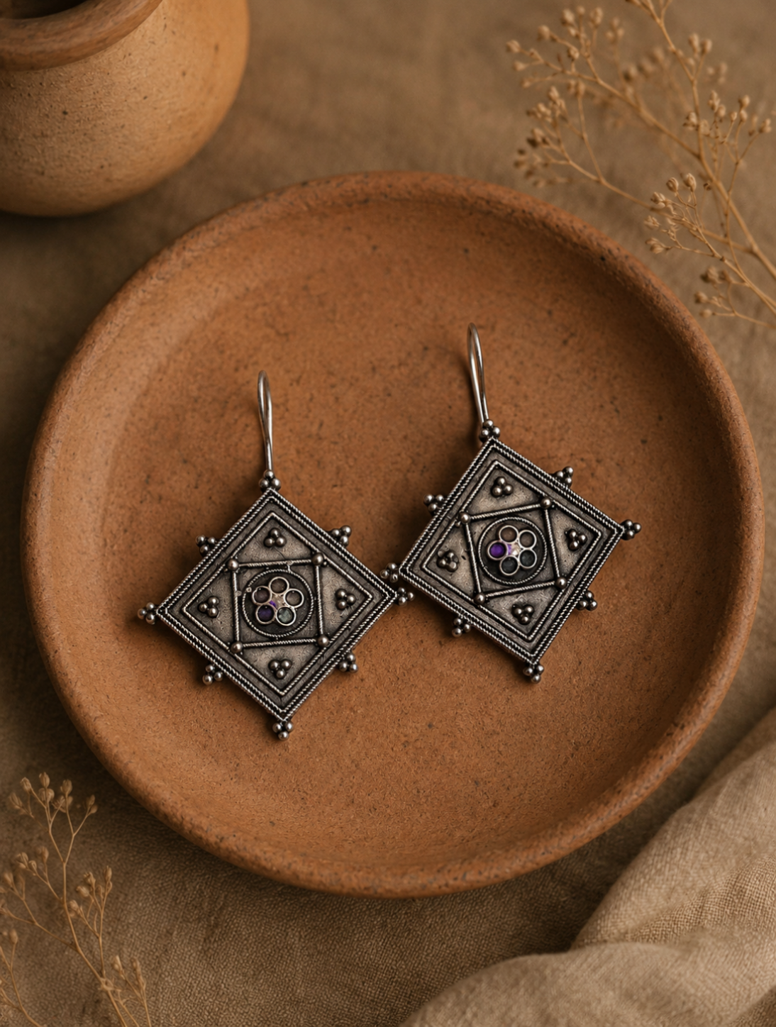 A pair of Jaji oxidized silver geometric earrings featuring a diamond shape with intricate filigree borders, raised metallic dots, and a small purple stone center, displayed on a clay plate.