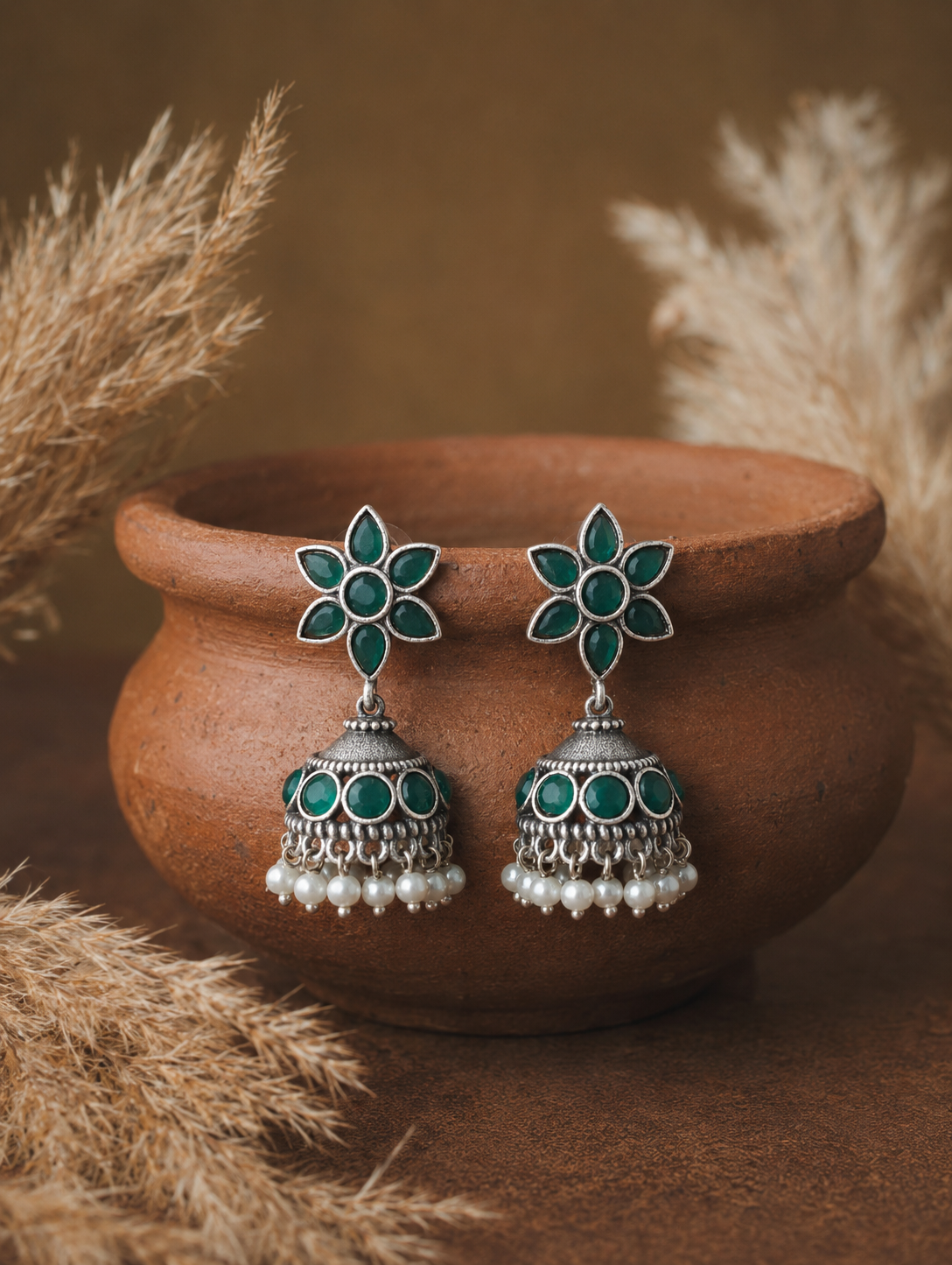 Floral earrings with green gemstones on a brown pot with dried plants.