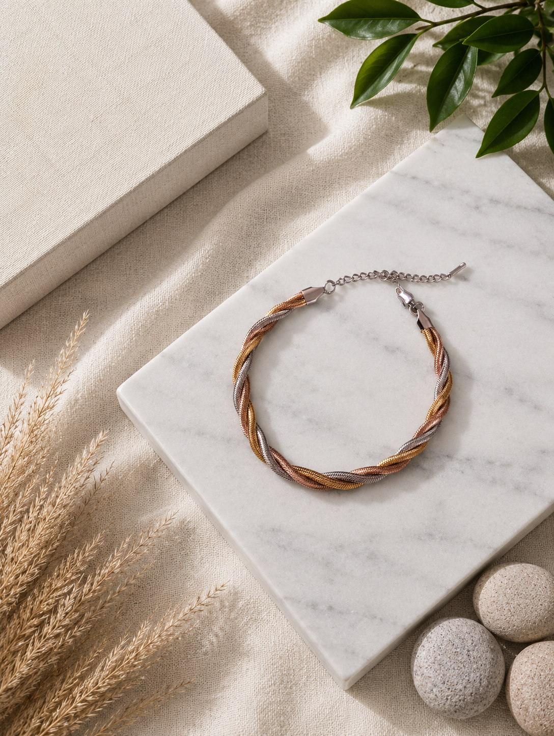 A top-down product photograph of a tricolour anklet arranged neatly on a square white marble slab. The anklet consists of three braided metallic snake chains in yellow gold, rose gold, and silver tones, finished with a silver link extender chain and clasp. The aesthetic background features textured off-white linen, dried pampas grass stalks, smooth river stones, and fresh green leaves.