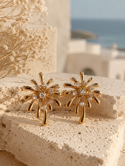 A flat-lay product photograph of a pair of oversized gold starburst stud earrings from the Diamond Pop collection. The earrings are displayed upright on porous cream-colored travertine stone blocks under warm, natural sunlight, showcasing a highly polished yellow-gold firework-like design adorned with scattered, sparkling clear crystals. The background features delicate dried white flowers and a soft, blurred view of a seaside architectural backdrop. As seen in GE012_Diamond POP_499.png.