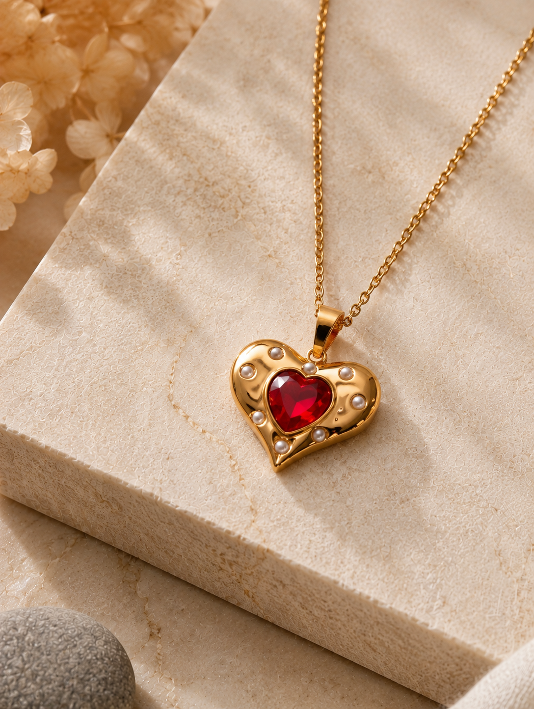 An elegant gold necklace with a red heart pendant and embedded crystals, displayed on a textured beige stone slab. The background features a minimalist, warm-toned aesthetic with cream ceramic vases, dried white hydrangea flowers, smooth gray river stones, and a soft white cloth, all illuminated by gentle sunlight and soft shadows.