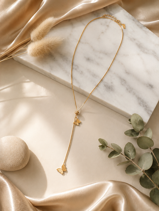 A gold lariat-style necklace featuring two delicate butterfly charms attached to a drop chain with an adjustable sliding bead. In GN007_butterfly chain.png, the necklace is displayed on a long, veined white marble block. The luxury flat-lay arrangement is framed by shimmering, draped champagne silk fabric, a smooth cream river stone, fluffy dried bunny tail grass, and a sprig of green eucalyptus under warm, bright lighting.
