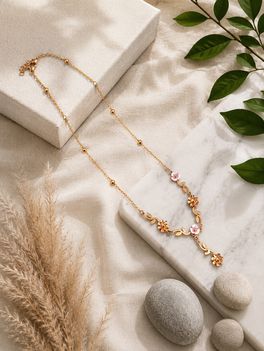 A close-up lifestyle shot in GN009_asymmetric floral chain_a.png showing a woman wearing a delicate, asymmetric gold necklace. The jewelry features an organic vine design decorated with small golden leaves, gold wildflowers, and white pearlescent blossoms, finished with a central dangling flower drop. The woman is wearing a ribbed, taupe off-the-shoulder top, and the scene is illuminated by soft, warm studio lighting that highlights her neckline and the necklace's intricate details.