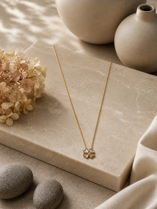 A delicate gold chain necklace with a four-leaf clover pendant displayed on a neutral beige fabric mannequin bust. The pendant in GN011_cute leaf chain.png features three heart-shaped leaves made of soft translucent stone, while the fourth leaf is filled with small sparkling crystals. The warm-toned, minimalist background includes draped soft fabric and a beige ceramic vase filled with dried pampas grass.