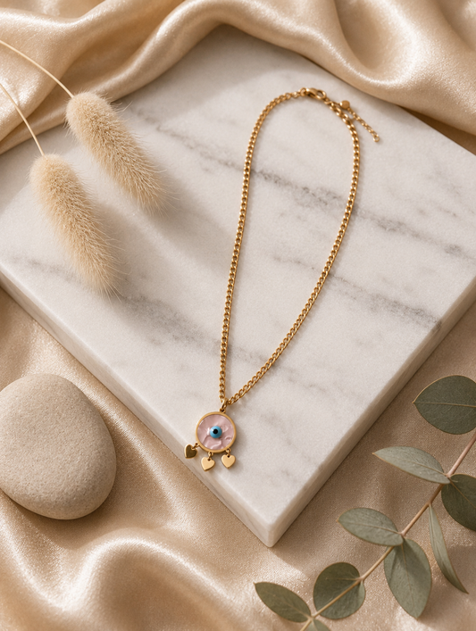 > **Alt Text:** A collection of product and lifestyle images featuring a gold-plated curb chain necklace with a circular evil eye pendant. The pendant showcases a pearlescent marbled inlay face, a vibrant cerulean blue and black eye at the center, and three dainty, polished gold heart charms dangling from its base. The necklace is displayed both as a flat-lay on a white marble slab with neutral aesthetic props—like a smooth river stone, dried grass, and draped champagne silk—and worn elegantly on a model's