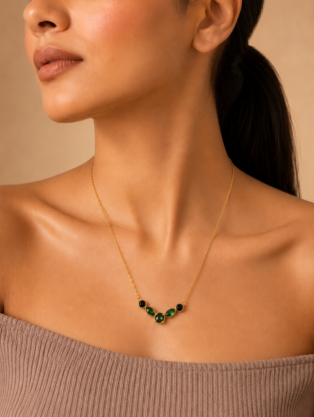 A product photography collage featuring a delicate gold-toned necklace with a curved station pendant of five faceted gemstones in rich emerald green and deep black. The first view shows a close-up of the necklace resting on a white marble slab with smooth stones and textured linen. The second shows the entire length of the gold link chain in a styled flat-lay next to a gift box and tropical leaves. The third shows the necklace being worn by a model, resting elegantly on her collarbone with a taupe off-the-s