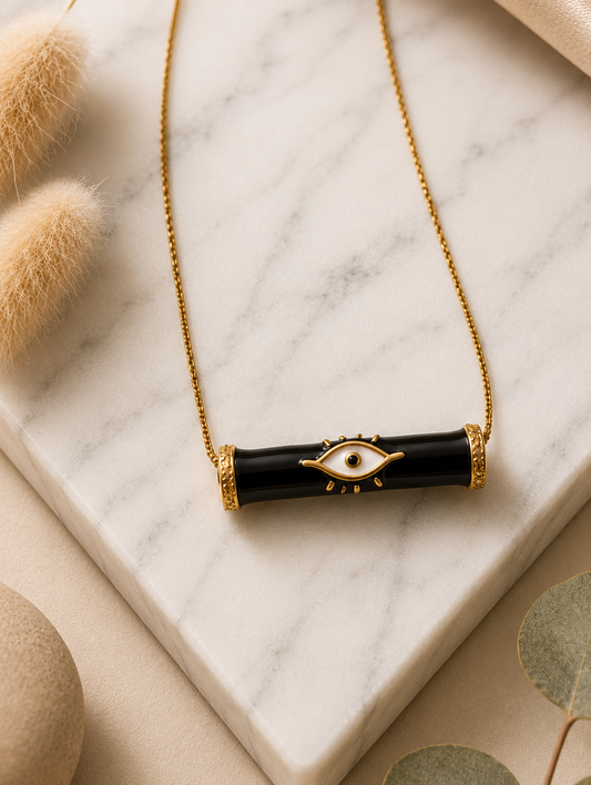 A product photography display showcasing a modern gold-toned necklace with a horizontal black enamel bar evil eye pendant. The collection includes a styled flat-lay view on a marble slab surrounded by champagne satin and dried botanicals, a display view of the necklace resting on a cream velvet jewelry mannequin, and a macro close-up highlighting the crisp white, black, and gold details of the central eye motif and its textured end caps.