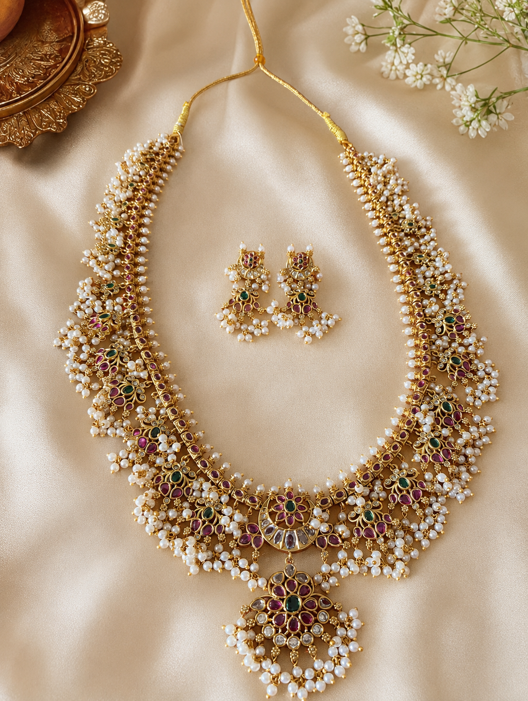 Flat lay view of the Revathy Long Guttapusalu necklace and its matching earrings laid gracefully on a shimmering cream fabric backdrop next to decorative gold plates Flat lay view of the Revathy Long Guttapusalu necklace and its matching earrings laid gracefully on a shimmering cream fabric backdrop next to decorative gold plates and white flowers.and white flowers.