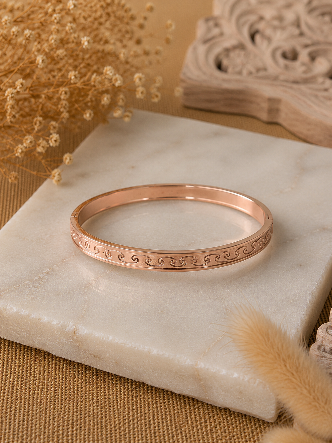 A flat-lay product photograph of a sleek rose-gold kada bracelet resting flat on a square white marble slab. The rigid bangle features an elegantly engraved continuous ocean wave and scroll motif along its outer band, framed by polished edges. The background is styled with dried baby's breath flowers, neutral tan woven textiles, and a carved wooden accent. As seen in KD024_rose gold kada_a.png.