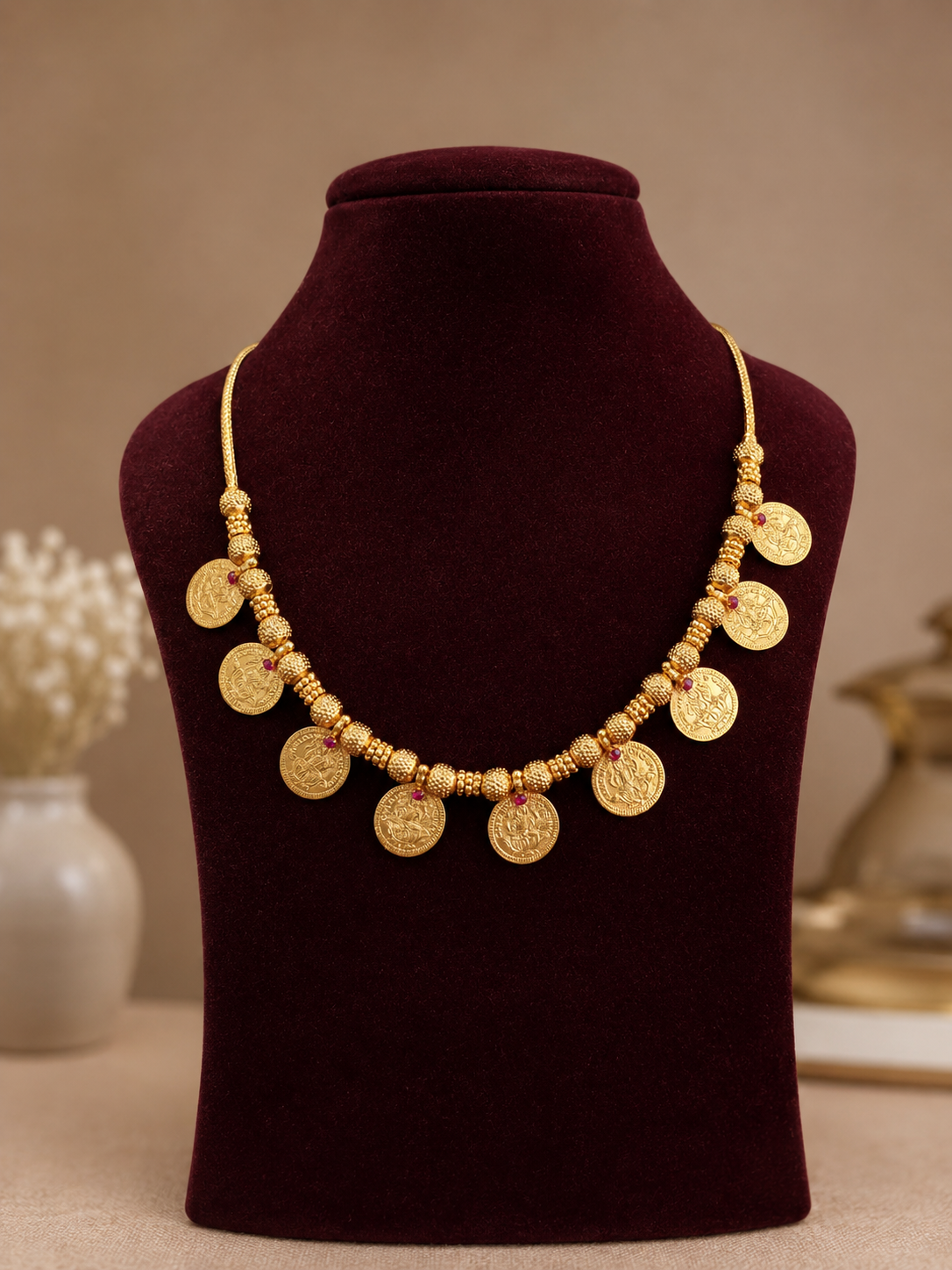 The Anagha traditional gold Kasu Mala necklace featuring 9 dangling Lakshmi coins spaced along a detailed gold bead chain with small red stone accents, displayed on a dark red velvet mannequin.