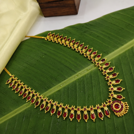 A traditional Kerala Nagapadam necklace featuring vibrant ruby red stones set in gold, displayed flat on a fresh green banana leaf next to a cream-colored fabric.