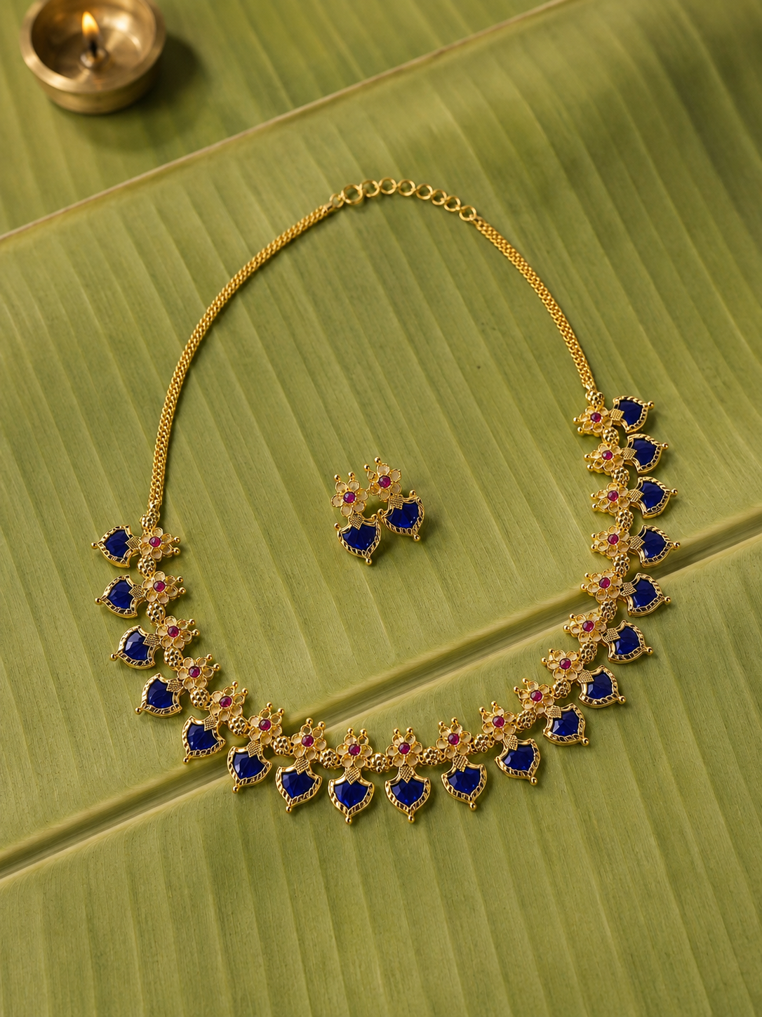 A royal blue stone variant of the traditional Kerala Palakka necklace and matching stud earrings set, displayed flat on a green banana leaf next to a lit brass oil lamp.