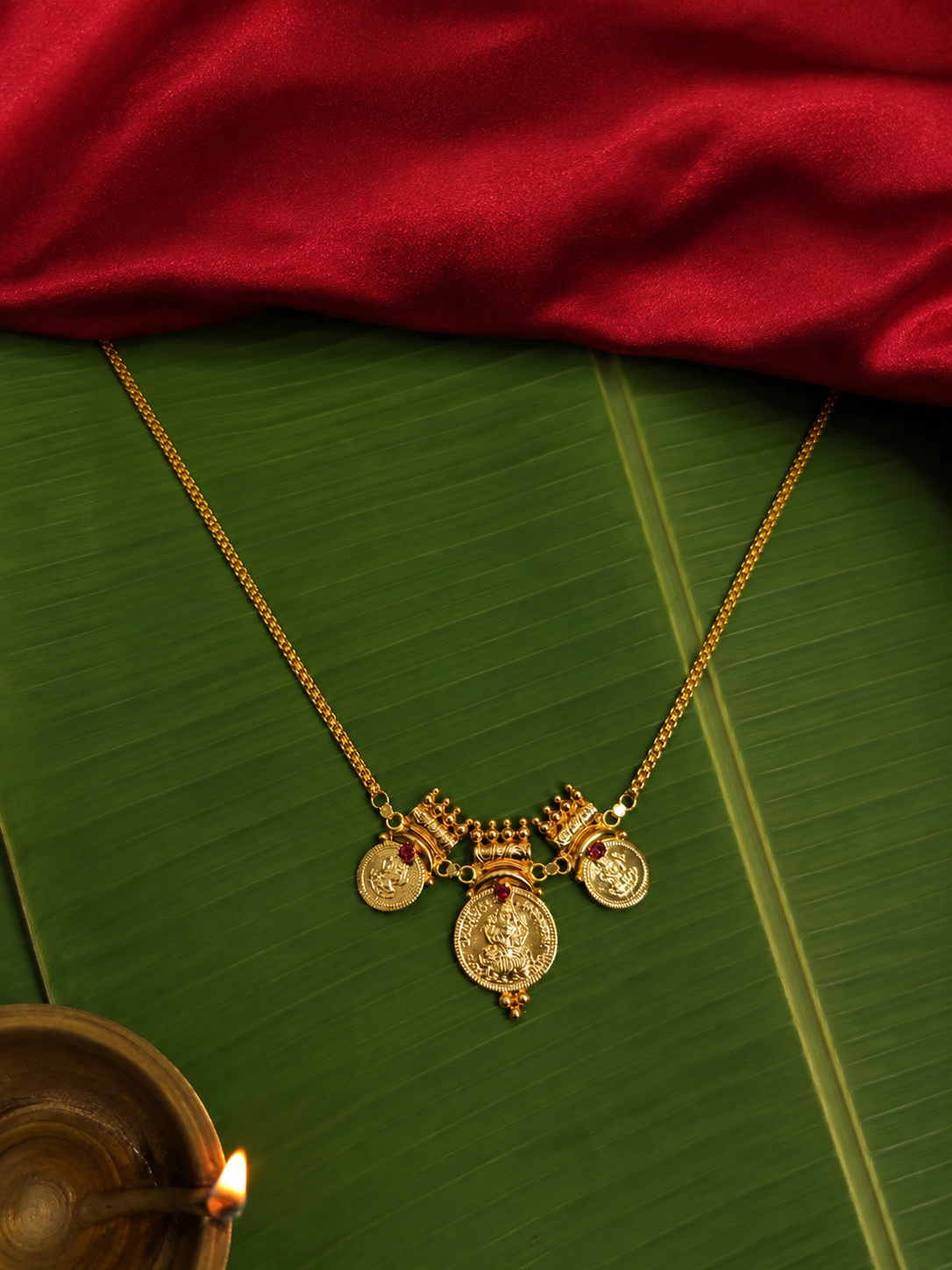 A delicate traditional gold chain necklace featuring three Goddess Lakshmi coin pendants accented with tiny pink rubies, placed flat on a vibrant green banana leaf below a rich red silk fabric.