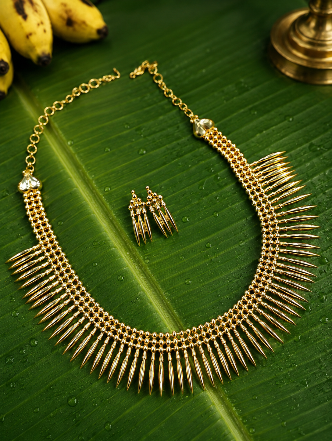 Flat lay view of an all-gold traditional Kerala Mullu Mottu Haram necklace and matching spike-shaped drop earrings set, arranged beautifully on a wet green banana leaf with fresh water droplets, a gold oil lamp, and yellow bananas in the background.