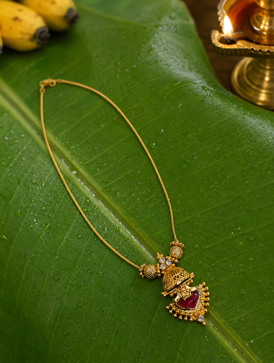 Elegant traditional gold necklace with a detailed Jhumka pendant accented by a rich red ruby-colored gemstone, placed flat on a wet banana leaf background.