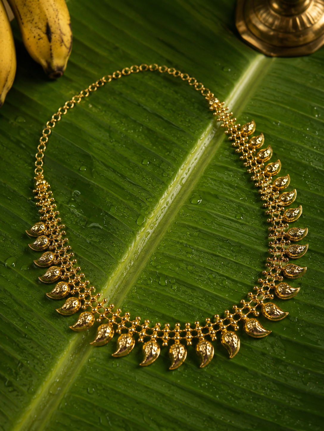 A traditional South Indian gold-toned Manga Mala necklace featuring mango-shaped paisley motifs and delicate beadwork, displayed elegantly on a wet, vibrant green banana leaf alongside yellow bananas and a brass vilakku base.