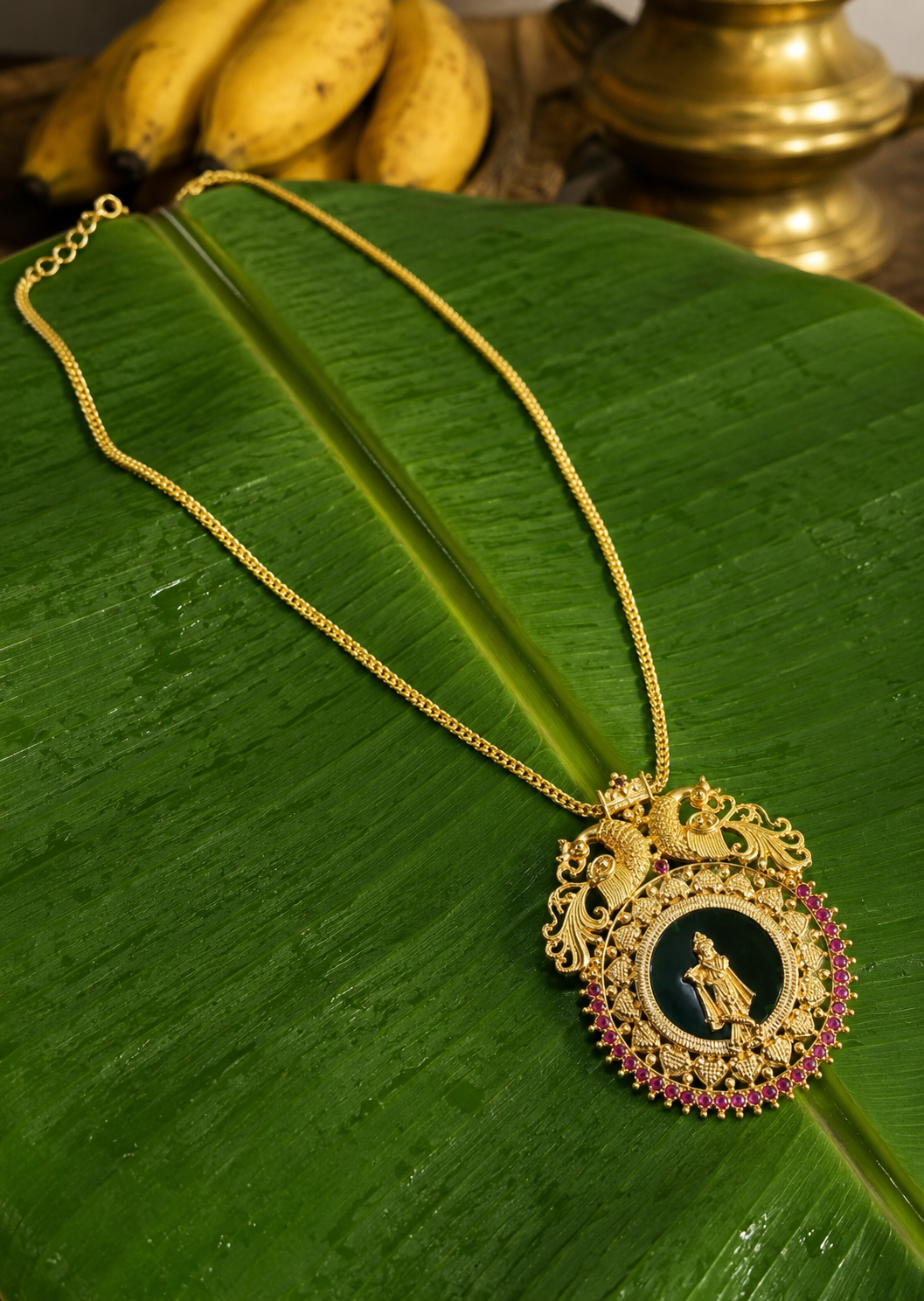 Top-down view of the Krishnaaa heritage pendant necklace featuring a dark forest green enamel center with the golden Lord Krishna icon, dual peacock filigree, and a ruby stone outline, elegantly displayed on a wet banana leaf.