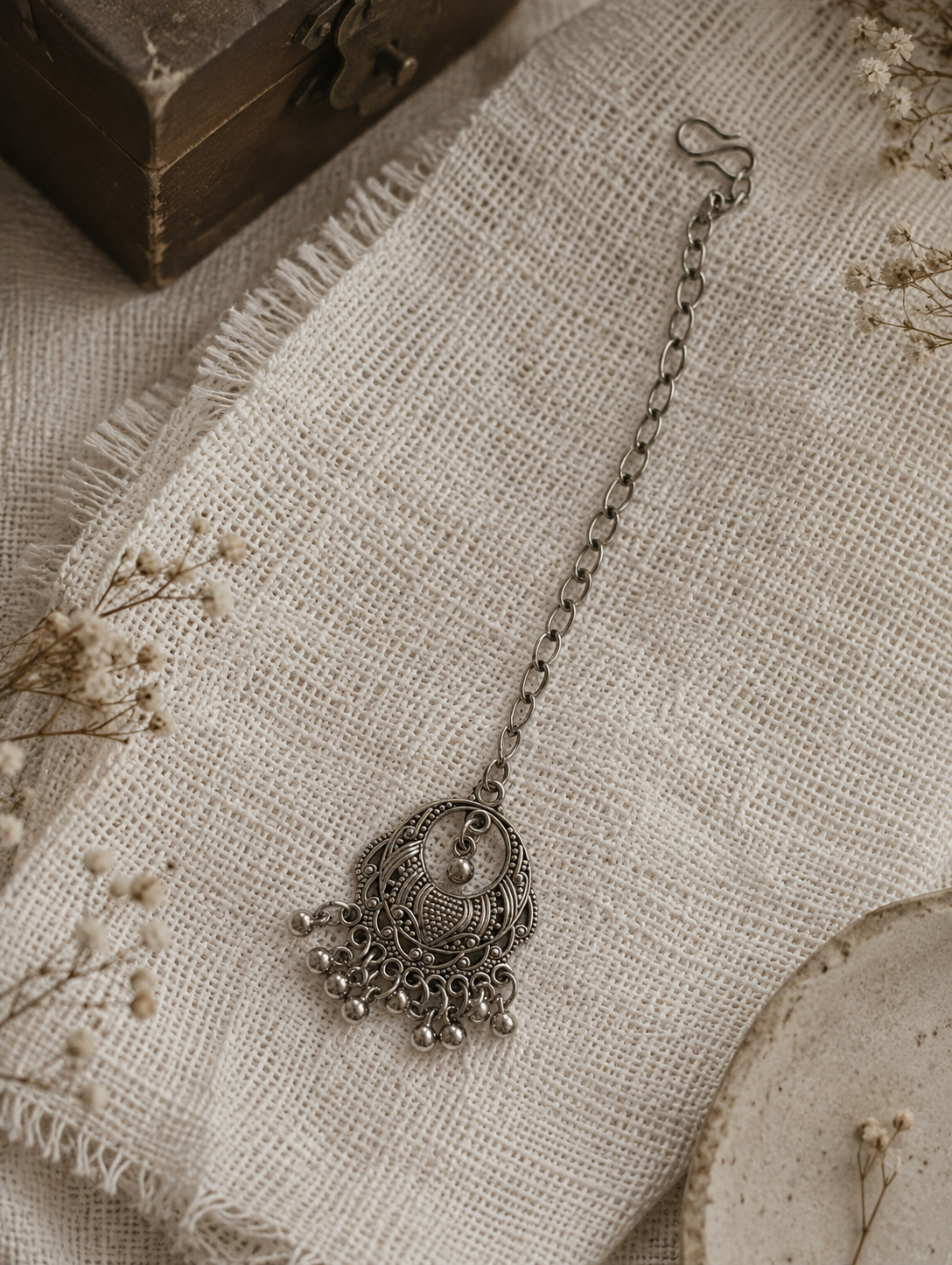 An overhead flat-lay shot of an oxidised silver maang tikka from the Dhanu collection, featuring an ornate crescent-shaped medallion with fine textured engravings and a fringe of tiny metallic ghungroo balls, attached to a simple metal link chain on a textured off-white linen cloth.