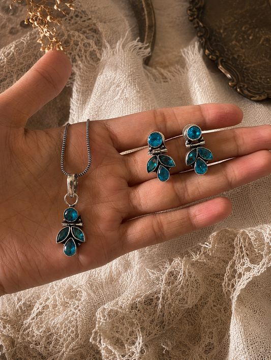 An oxidized silver jewelry set displayed on a hand, featuring a delicate chain necklace with a blue crystal leaf-shaped pendant and matching leaf-motif dangle earrings.