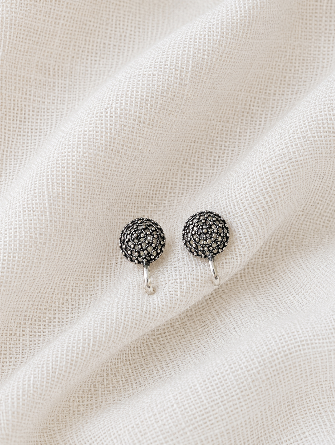 A close-up product shot of a pair of small, circular oxidized silver clip-on nose pins. Each nose pin features dense, textured rava metallic beadwork arranged in concentric patterns, displayed resting against a soft, textured white fabric backdrop.