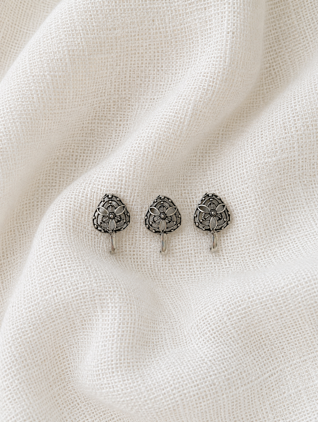 A close-up product photograph of three small, teardrop or leaf-shaped clip-on nose pins made of oxidized silver. Each nose pin is embellished with a delicate filigree pattern forming a central four-petal flower motif inside a textured, pointed metallic border, neatly arranged on a wrinkled white fabric background.