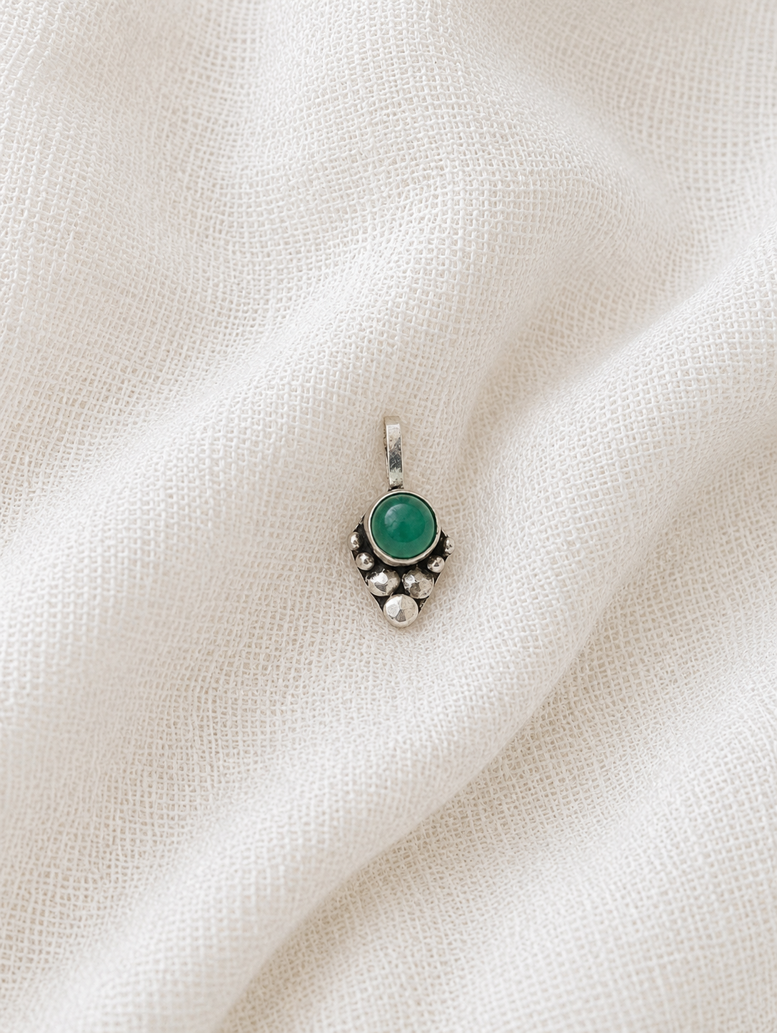 A high-angle product photograph of a single clip-on nose pin made of oxidized silver. The piece features a smooth, round green gemstone cabochon at the top, accented underneath by a descending cluster of small, textured metallic beads that form a tapered, triangular point, resting flat against a soft, crinkled white fabric background.