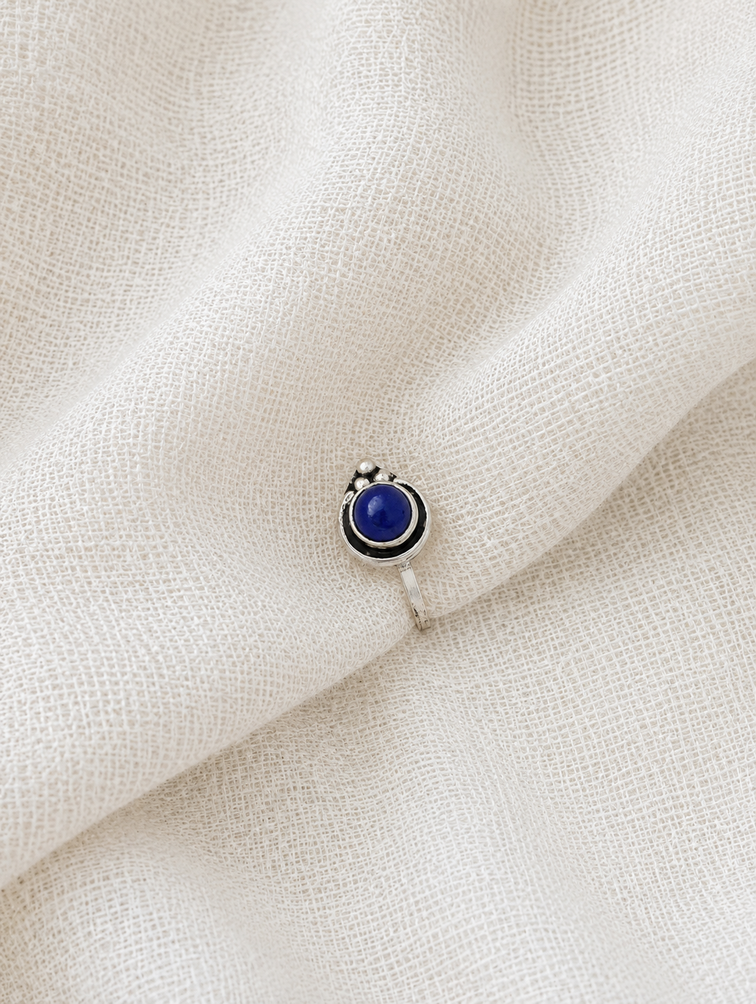 A small silver teardrop-shaped nose pin with a prominent round, dark blue lapis-like stone center and silver bead detailing, resting on a draped, off-white textured fabric background