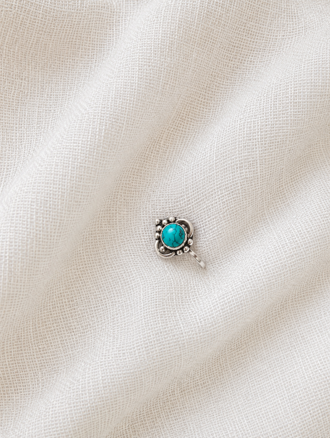 A small, ornate oxidized silver nose pin featuring a round turquoise gemstone center with delicate silver beadwork and curved accents, displayed on a textured off-white fabric background